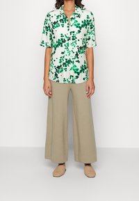 Blusa a maniche corte con un motivo floreale verde su sfondo bianco, abbinata a pantaloni larghi beige e scarpe slip-on in tan intrecciate.