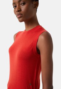 Czerwony sweter bez rękawów wykonany z miękkiego, gładkiego dzianiny. Posiada wysoki kołnierz oraz prążkowane wykończenia pod pachami, a na dole znajduje się mała metka z logo.