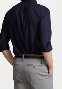 Camisa de botón azul marino con mangas dobladas, combinada con pantalones gris claro y un cinturón trenzado marrón. Presenta un pliegue en la parte posterior y un detalle de logo.