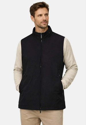 Uomo che indossa un gilet nero con zip sopra una camicia beige a maniche lunghe, con le mani in tasca, che guarda leggermente di lato.