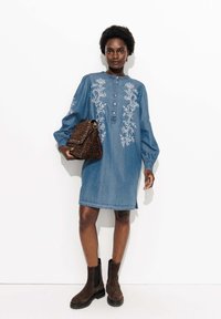 Robe en denim à manches longues, avec un design de broderie florale blanche et des détails de boutons. Bottines en daim marron à la cheville et un sac à main à imprimé léopard.