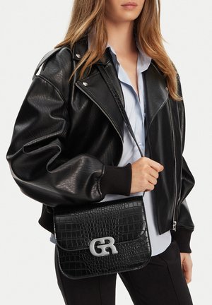 Mujer con chaqueta de cuero negra y camisa azul claro, sosteniendo un bolso cruzado negro con textura de cocodrilo y hebilla plateada con iniciales.