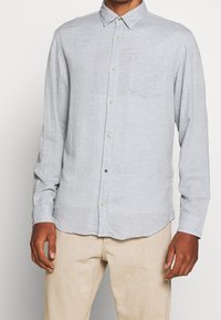 Camicia azzurro chiaro con tessuto strutturato e motivo, maniche lunghe, taschino sul petto e bottoni beige. Indossata con pantaloni beige.