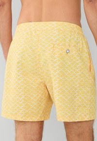 Žute kupaćice sa ponovljenim bijelim uzorkom školjki, elastičnom naramenicom i stražnjim džepom s kopčanjem na gumb.