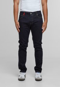 Sötétkék, slim fit farmernadrág öt zsebbel, narancssárga varrással és egy jellegzetes piros kiegészítővel az első zsebben.