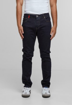 Man in donkere blauwe slim-fit jeans, witte sneakers met zwarte strepen, en een wit mouwloos shirt, staand tegen een effen achtergrond.