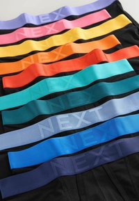 Pile de boxers noirs avec des ceintures élastiques en violet, rose, jaune, orange, rouge, sarcelle, gris clair, bleu et marine, chacun marqué "NEXT".