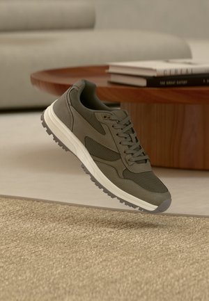 Oliwkowo-zielony sneaker z białą podeszwą unoszący się nad beżowym, plecionym dywanem w salonie z drewnianym stolikiem kawowym i ułożonymi książkami.