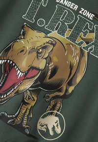 Maglietta verde con una grande grafica dettagliata di un T. rex in marrone e beige, con denti e artigli pronunciati e una silhouette circolare del dinosauro.
