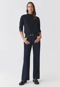 Maglione lavorato a maglia blu navy con dettagli ai polsini, abbinato a jeans scuri a vita alta e una cintura nera. Il modello indossa scarpe nere lucide.