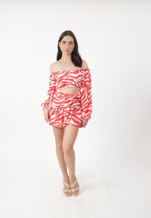 Frau in korallfarbenem und weißem Zebra-Print Off-Shoulder Crop-Top und passendem Wickelrock, beige Absatzsandalen, vor weißem Hintergrund stehend.