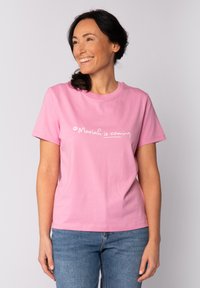 Glunderende vrouw met donker haar in een paardenstaart draagt een roze T-shirt met de tekst "Mariah komt eraan." en blauwe jeans, staande tegen een effen achtergrond.