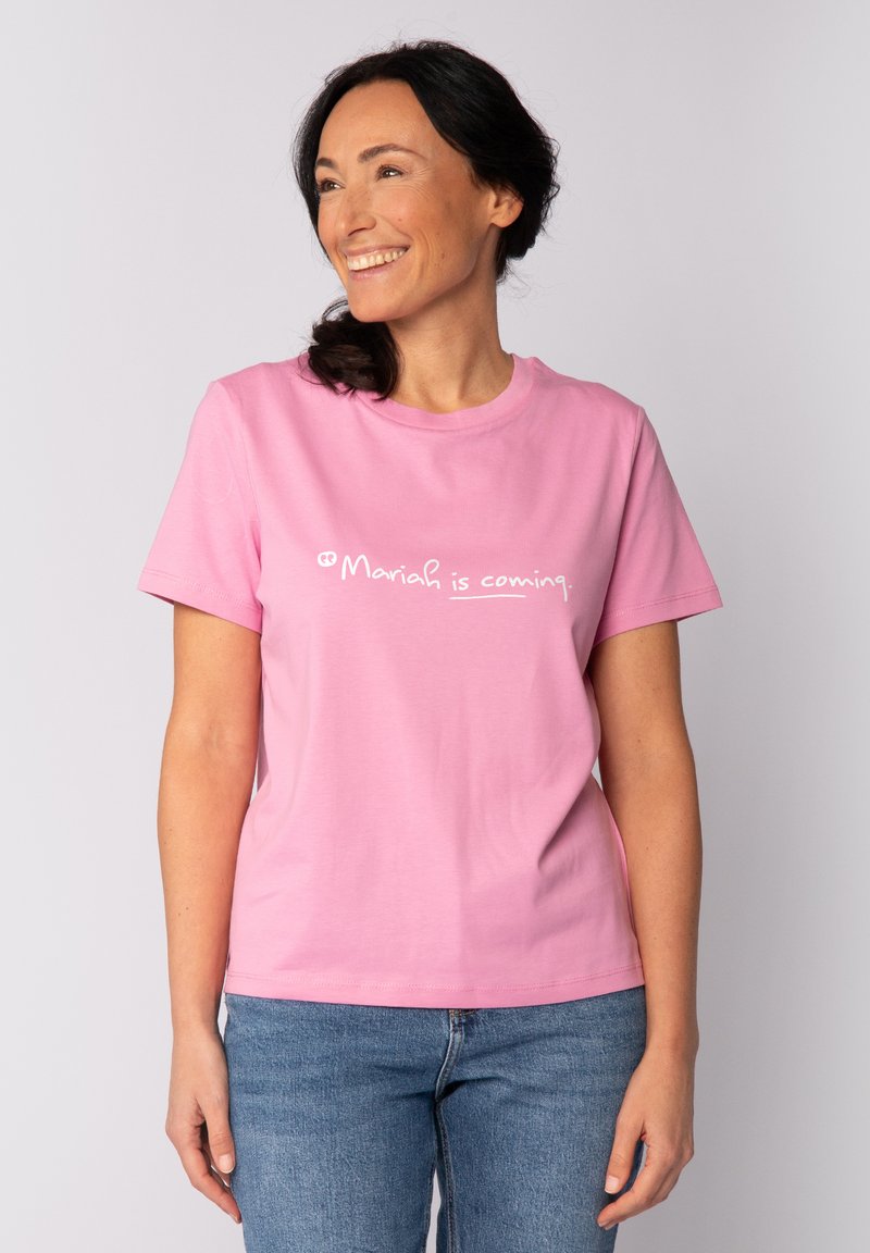 Glunderende vrouw met donker haar in een paardenstaart draagt een roze T-shirt met de tekst "Mariah komt eraan." en blauwe jeans, staande tegen een effen achtergrond.