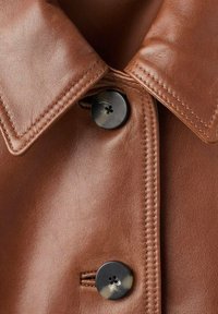 Veste en cuir marron avec un col pointu, des coutures visibles et une fermeture à bouton avec un détail de bouton circulaire noir.