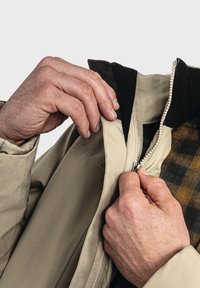 Hände, die eine beige Jacke über ein kariertes Hemd zippen, mit Fokus auf dem Reißverschluss und den Details des Jackenkragens.