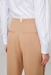 Beżowe spodnie stosowane pokazane z tyłu, charakteryzujące się gładką fakturą, dwoma tylnymi kieszeniami oraz średnim stanem, w zestawieniu z niebieską koszulą w paski.