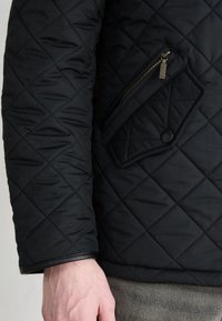 Μαύρο επενδυμένο μπουφάν με διαγώνια τσέπη με φερμουάρ, δερμάτινη επένδυση στα μανίκια και υφή με διακοσμητές.