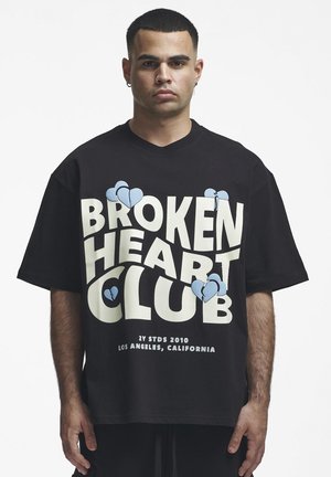 Férfi fekete, túlméretezett pólót visel az elülső részen "Broken Heart Club" felirattal és kék tört szív illusztrációkkal.