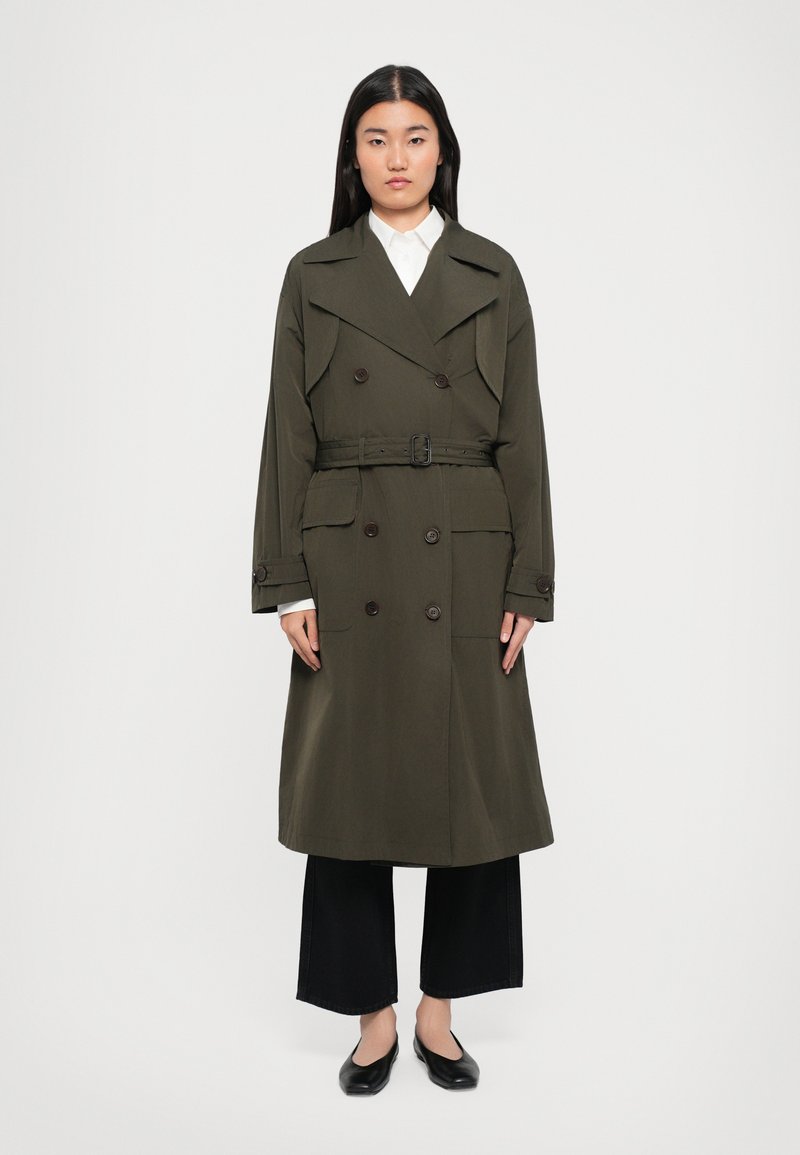 Olivengrüner Trenchcoat mit doppelreihigen Knöpfen, breitem Kragen, taillierter Taille und Vordertaschen; kombiniert mit schwarzen weit geschnittenen Hosen und Ballerinas.