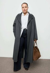 Cappotto di lana grigia con un ampio colletto, indossato sopra una camicia bianca e jeans svasati blu scuro. Borsa in camoscio marrone portata sul braccio destro.