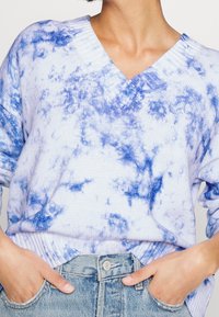 Ljust blå stickad tröja med V-ringning och texturerad tyg, med ett tie-dye-mönster i mörkare blå nyanser. Kallas med ljus tvättad denim.
