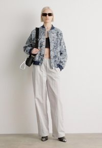 Veste bombardier à motif floral bleu marine, pantalon blanc ample et escarpins gris brillant. Sac noir avec chaîne en argent, lunettes de soleil noires élégantes.