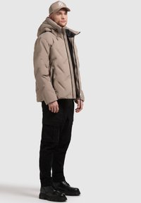 Giacca puffer beige con cappuccio, con zip frontale e due tasche laterali. Abbinata a pantaloni cargo neri e stivali neri con suola spessa.