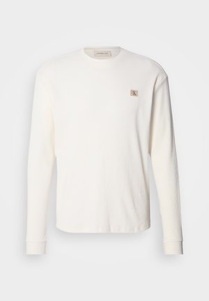 Λευκό ριμπ κοντομάνικο με στρογγυλή λαιμόκοψη, με ετικέτα Calvin Klein Jeans στο στήθος και μανσέτες στα μανίκια, παρουσιασμένο σε μονόχρωμο φόντο.