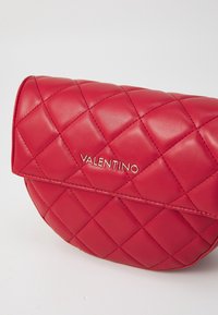 Quiltad röd läderväska med en rundad form, featuring en gyllene logotyp och en klaffstängning. Texturen är jämn med en glänsande yta.