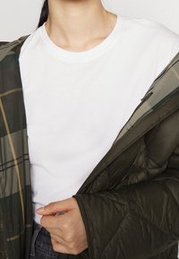 Zelená prošívaná péřová bunda s podšívkou s kostičkovaným vzorem, nošená přes jednobarevné bílé tričko s kulatým výstřihem. Detailní záběr na límec a ruku.