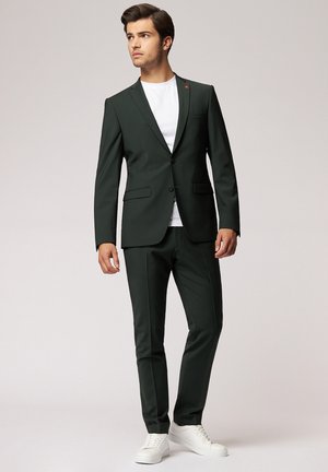 Young man in dark green suit with white shirt and white sneakers stands looking to the side against plain background.