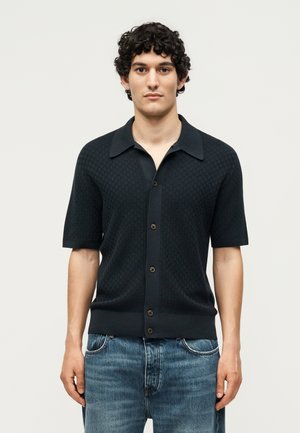 Jeune homme aux cheveux bouclés portant une chemise noire à manches courtes boutonnée et un jean bleu, debout devant un fond clair uni.