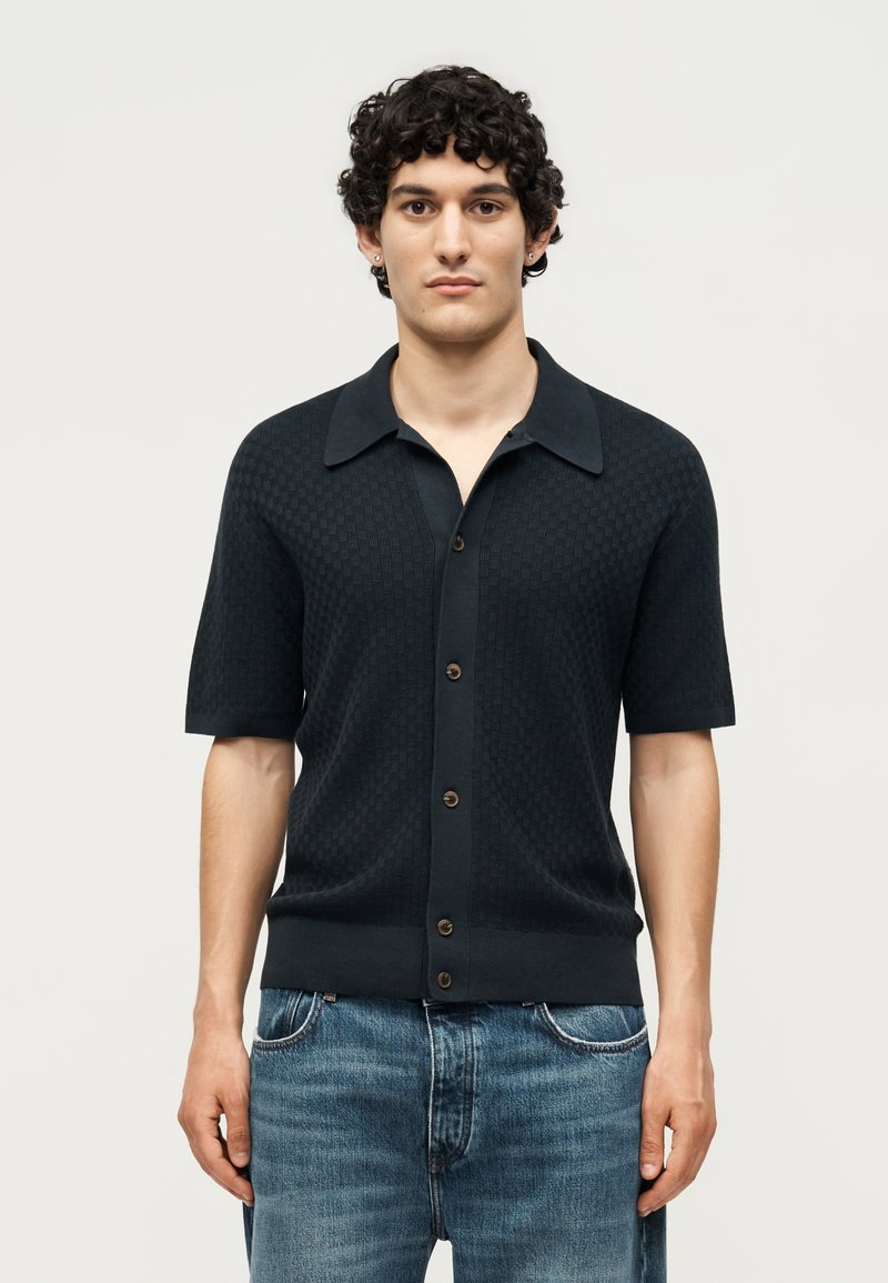 Νέος άνδρας με σγουρά μαλλιά, φορώντας μαύρο κοντομάνικο πουκάμισο με κουμπιά και μπλε τζιν, στέκεται μπροστά σε απλή φωτεινή φόντο.