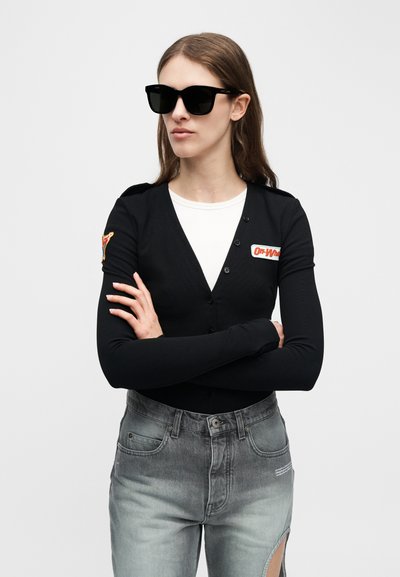 Femme portant un cardigan noir boutonné par-dessus un haut blanc, un jean gris fendu à la cuisse et de grandes lunettes de soleil noires, debout les bras croisés.