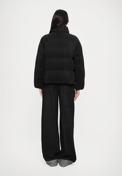 Black puffer jacket with vertical ribbed texture, high collar, and wide sleeves, paired with relaxed-fit black denim pants featuring gray cuffs.