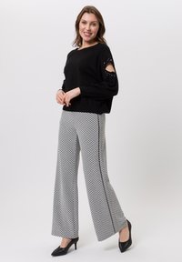 Lachende vrouw draagt een zwarte trui met uitgesneden mouwen en zwart-witte gestreepte broek met wijde pijpen, poseert met samengevouwen handen.