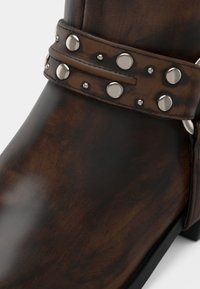 Καφέ δερμάτινο μποτάκι με διακοσμητική λουρίδα με καρφιά γύρω από τον αστράγαλο και χαμηλό μαύρο τακούνι, σε κοντινή λήψη.