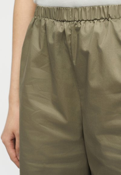 Povečava olivno zelenih hlač z elastičnim pasom, z naborki na tkanini in roko, ki počiva ob bokih.
