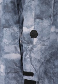 Tuulitakki teksturoidusta harmaasta kankaasta, jossa on pilvimäinen kuvio. Varustettu kuusikulmaisella logopatchilla ja säädettävillä tarranauhakasoilla.