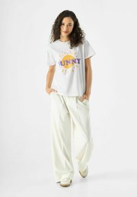 Weißes Baumwoll-T-Shirt mit einem bunten "SUNNY"-Grafikdesign und gelben Sonnenstrahlen. Kombiniert mit locker sitzenden, cremefarbenen Hosen und beigen Sneakers.