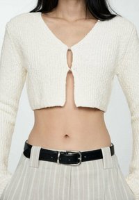 Pull blanc court à texture côtelée, col en V et détail œillet à l'avant. Associé à un pantalon taille haute à rayures claires et une ceinture noire.