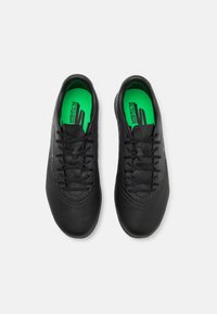 Chaussures de sport noires avec une tige texturée, un capuchon lisse et des lacets noirs. Les semelles intérieures sont d'un vert vif avec des détails contrastants.