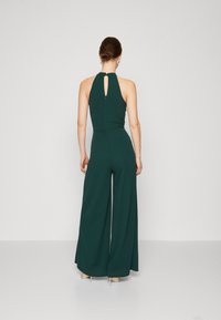 Grønt jumpsuit med halterneck, åpen nøkkelhullrygg og vide ben. Glatt stoff med tettsittende kropp og minimalt med maskinvaredetaljer.