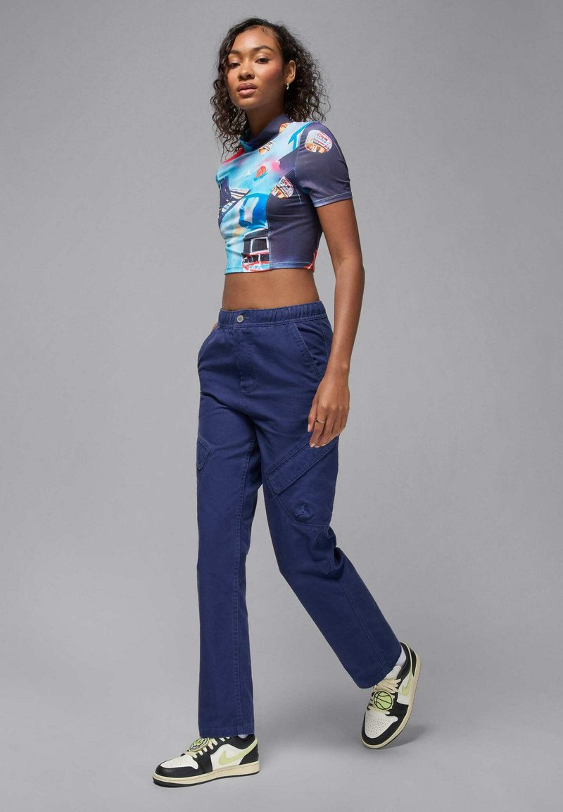 Graficzny crop top z niebieskimi i wzorzystymi detalami, zestawiony z granatowymi spodniami cargo; posiada elastyczną talię i wiele kieszeni. Czarno-białe sneakersy.