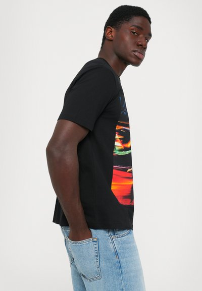 Side view of a man wearing a black graphic t-shirt and light blue jeans with hands in pockets against a white background.