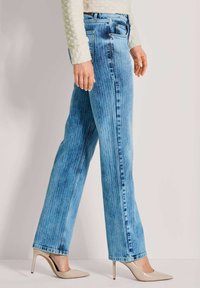 Vrouw in lichtblauwe rechttoe rechtaan jeans met verticale strepen, beige hoge hakken en een crèmekleurig gestructureerd langemouwen top, staande in zijaanzicht.