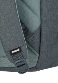 Cinghia di uno zaino in tessuto grigio con superficie testurizzata, caratterizzata da un logo Thule e hardware in plastica regolabile per il fissaggio.