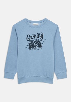 Sweat-shirt bleu clair en tissu doux, avec un motif graphique d'une manette de jeu et le texte "HARD CORE Gaming" imprimé en encre foncée.