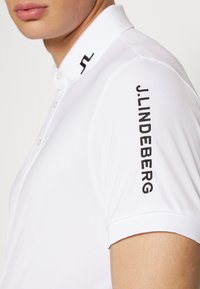 Camisa polo branca com colarinho clássico, apresentando uma abertura com botões e um logótipo bordado em preto "J.LINDEBERG" na manga direita.