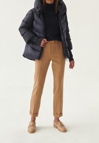 Puffatakki tummansinisessä, korkea kaulus, yhdistetty räätälöityihin kamelinvärisiin housuihin ja beigeihin loafereihin, joissa on kultaiset yksityiskohdat. Sileät pinnat.